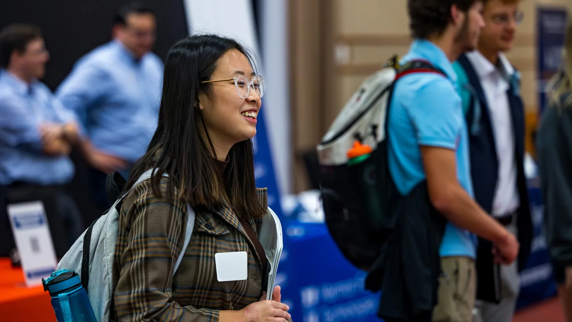A smiling young woman with long dark hair and clear framed glasses stands in profile in a hall with blue banners, wearing a plaid jacket and a gray backpack, holding papers against her chest, with a blank white name tag on her jacket and blurred people in blue shirts behind her.