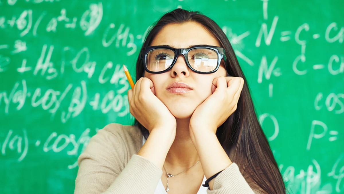 A young woman with long dark hair and black framed glasses rests her chin on both hands and looks upward with a pursed expression, holding a yellow pencil, in front of a green chalkboard covered in chalk equations.