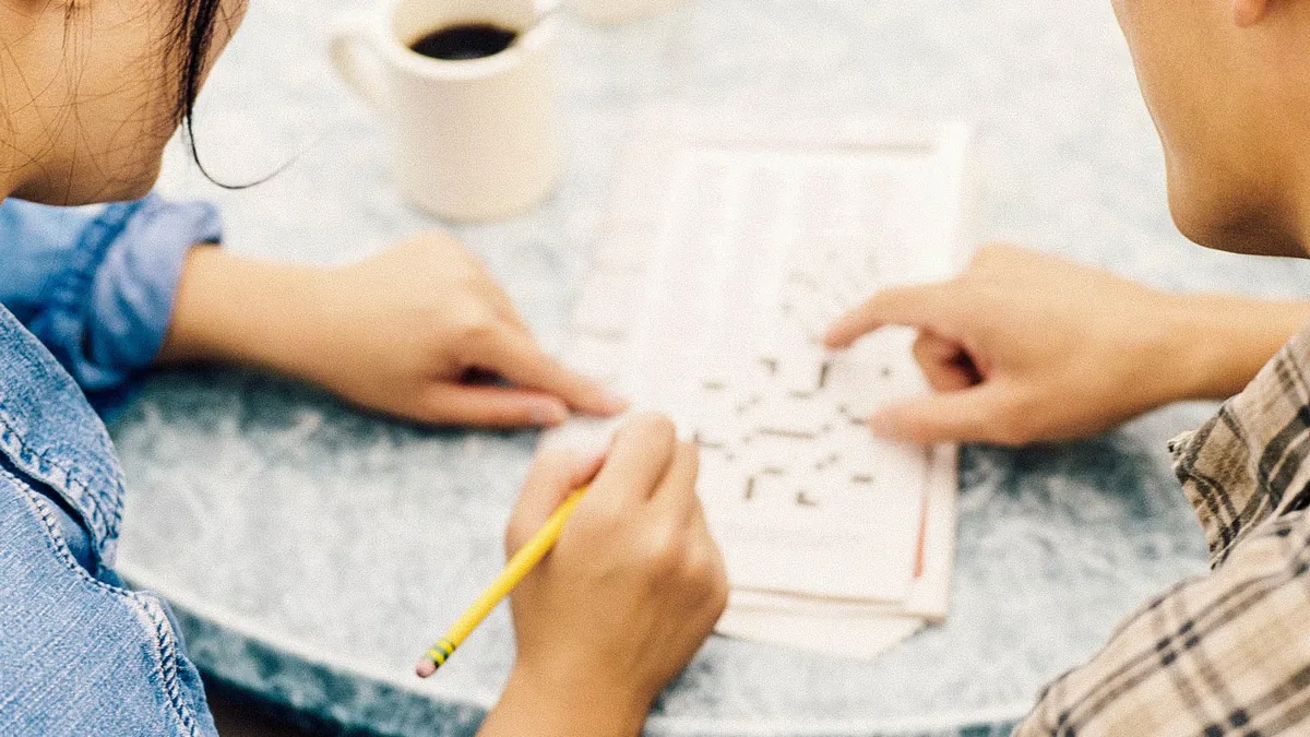 Two people sit close together at a marble topped table, one in a denim jacket holding a yellow pencil and the other in a plaid shirt, with their hands resting on a printed puzzle grid beside a white mug of coffee.