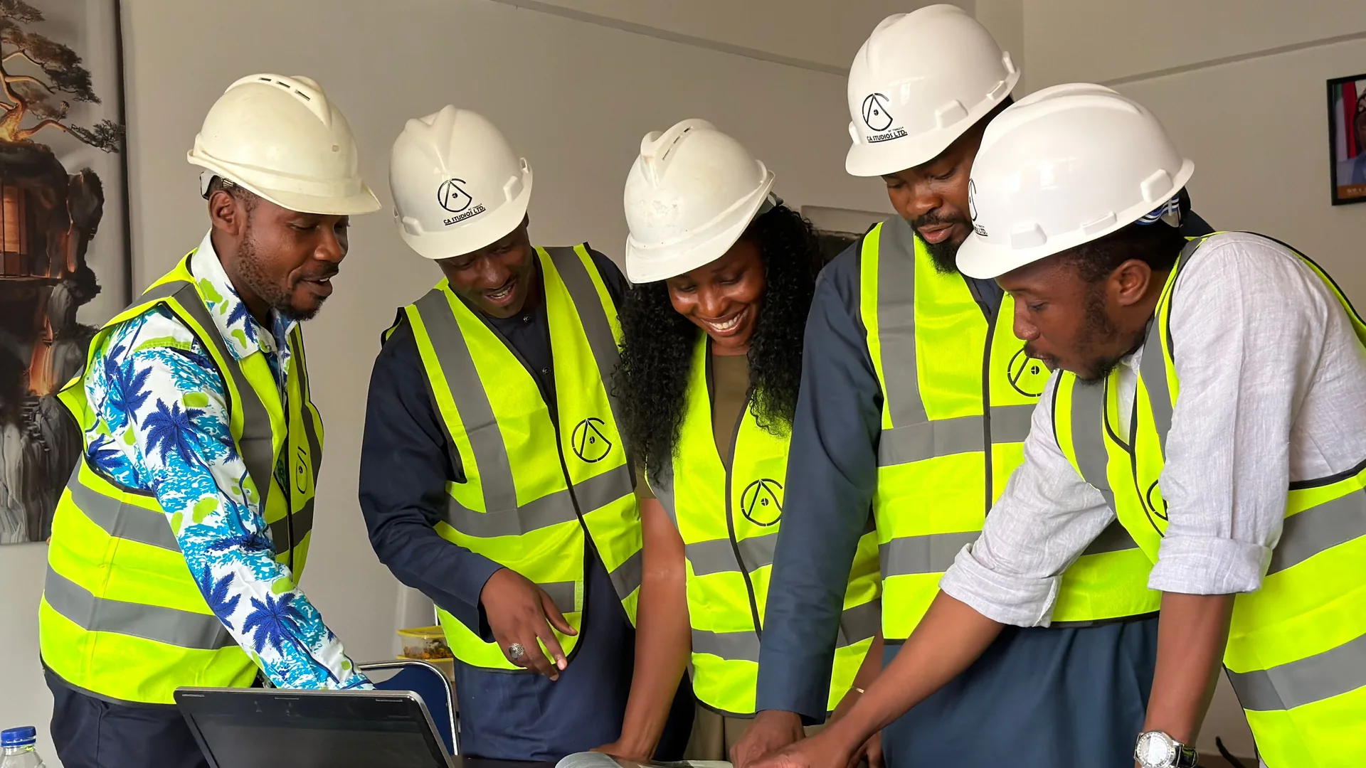 Five engineers in white hard hats and yellow safety vests gather around plans and a laptop.
