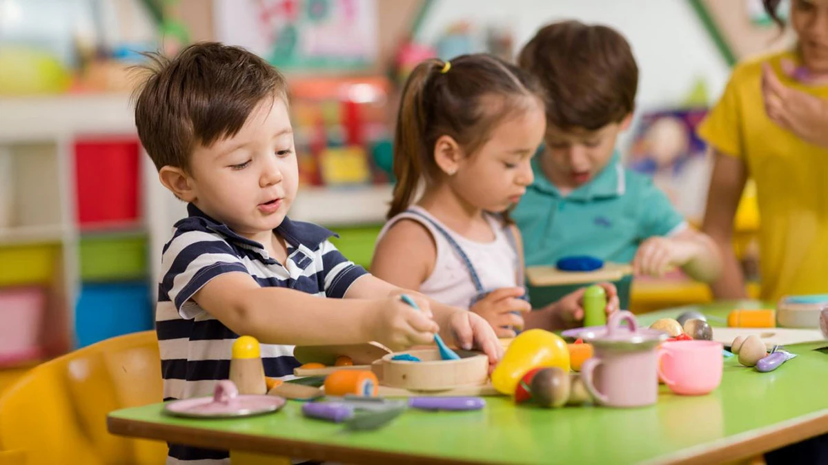 Three small children sit at a green table handling wooden toy food, cups and utensils, while an adult in a yellow top is partly visible at the right edge of the frame.