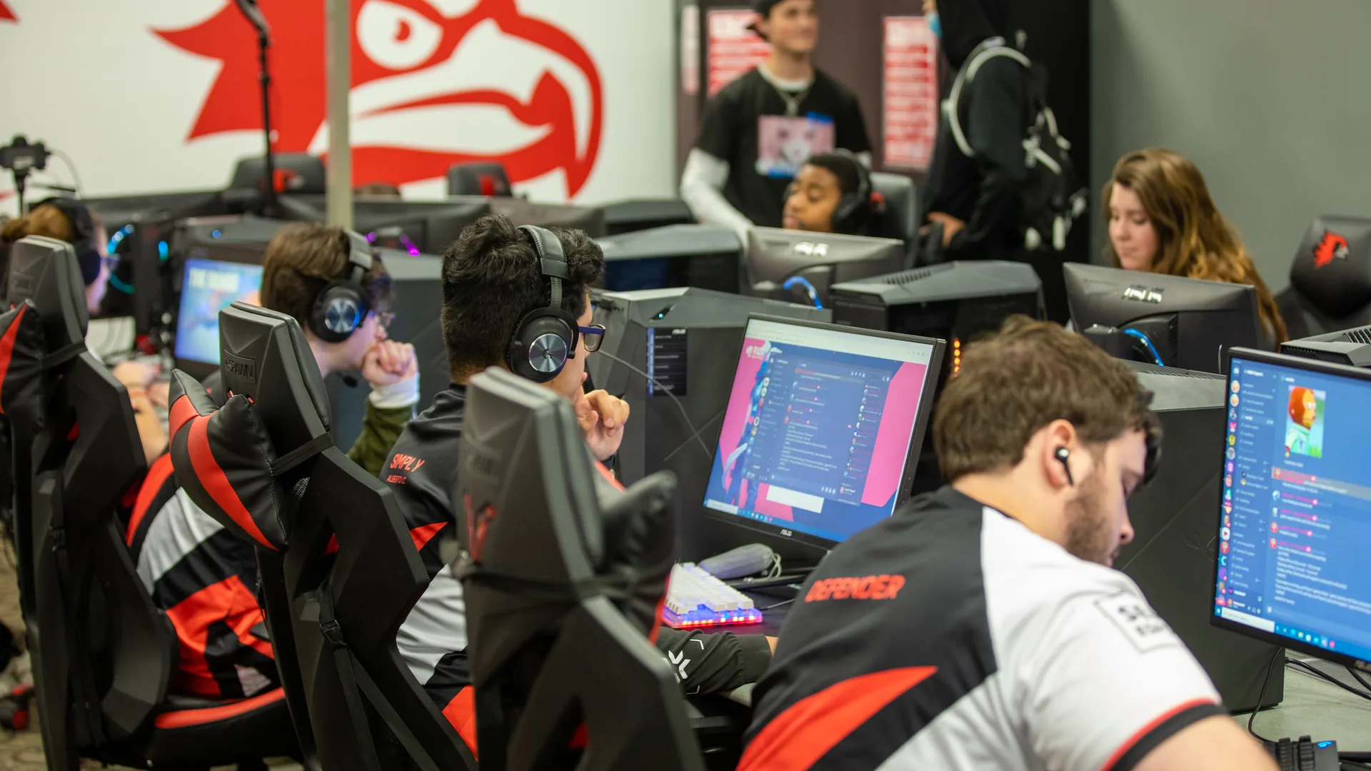 Players in team jerseys and headsets sit at rows of gaming computers in an esports room.