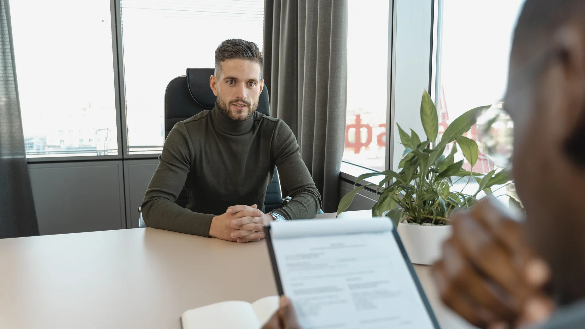 A man in a dark green sweater sits across a desk from a person holding a clipboard with a printed document.