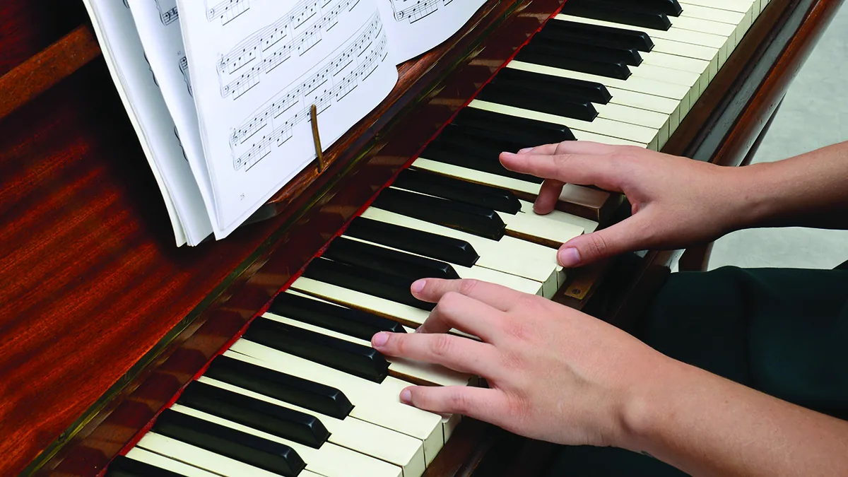 Two hands play an upright piano beneath an open book of sheet music.