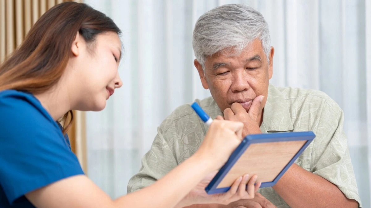 A woman in a blue top writes with a blue marker on a small framed board while an older man with gray hair rests his hand on his chin and looks down at the board, in a room with sheer white curtains.