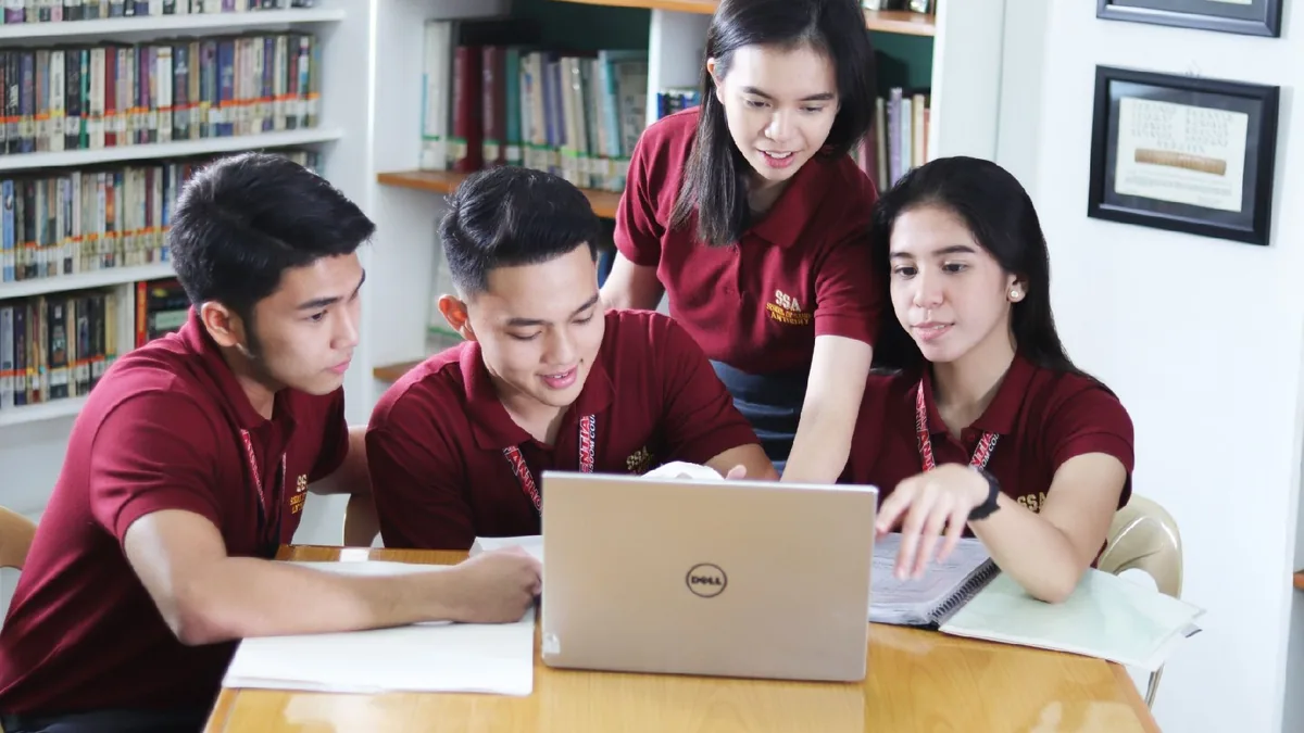 Four young people in maroon polo shirts with lanyards gather around a silver laptop on a wooden table in front of library shelves, one standing and leaning in toward the screen while another points at it.