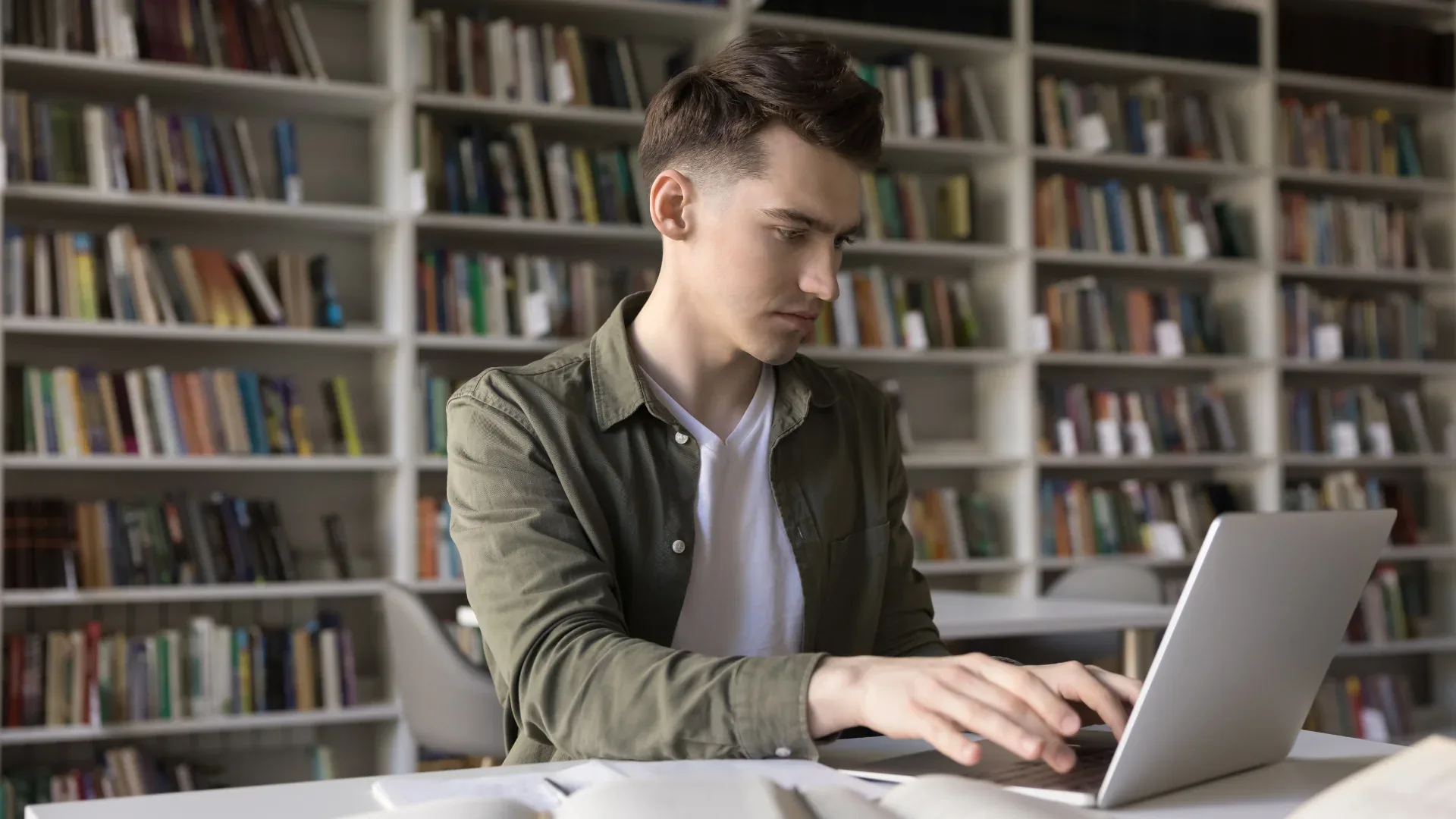 A young man types on a laptop at a table in front of tall library shelves.