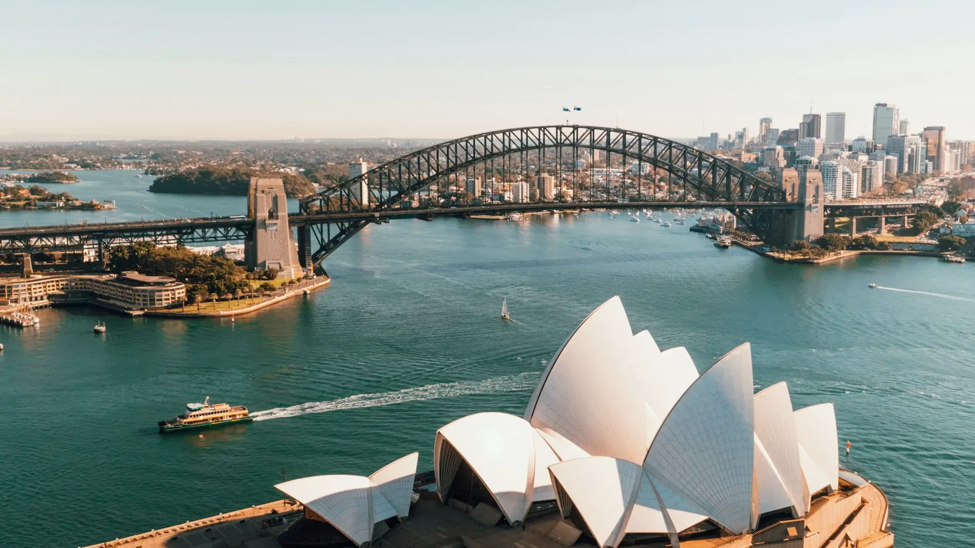 Aerial view of Sydney Harbour in daylight, with the white shells of the Sydney Opera House in the foreground, a ferry crossing the water and the Harbour Bridge arching across the middle distance beside city towers.