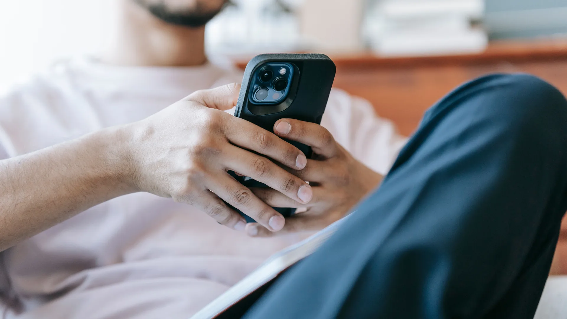 A man in a light T-shirt holds a smartphone in a black case with both hands, one knee raised, seated indoors.