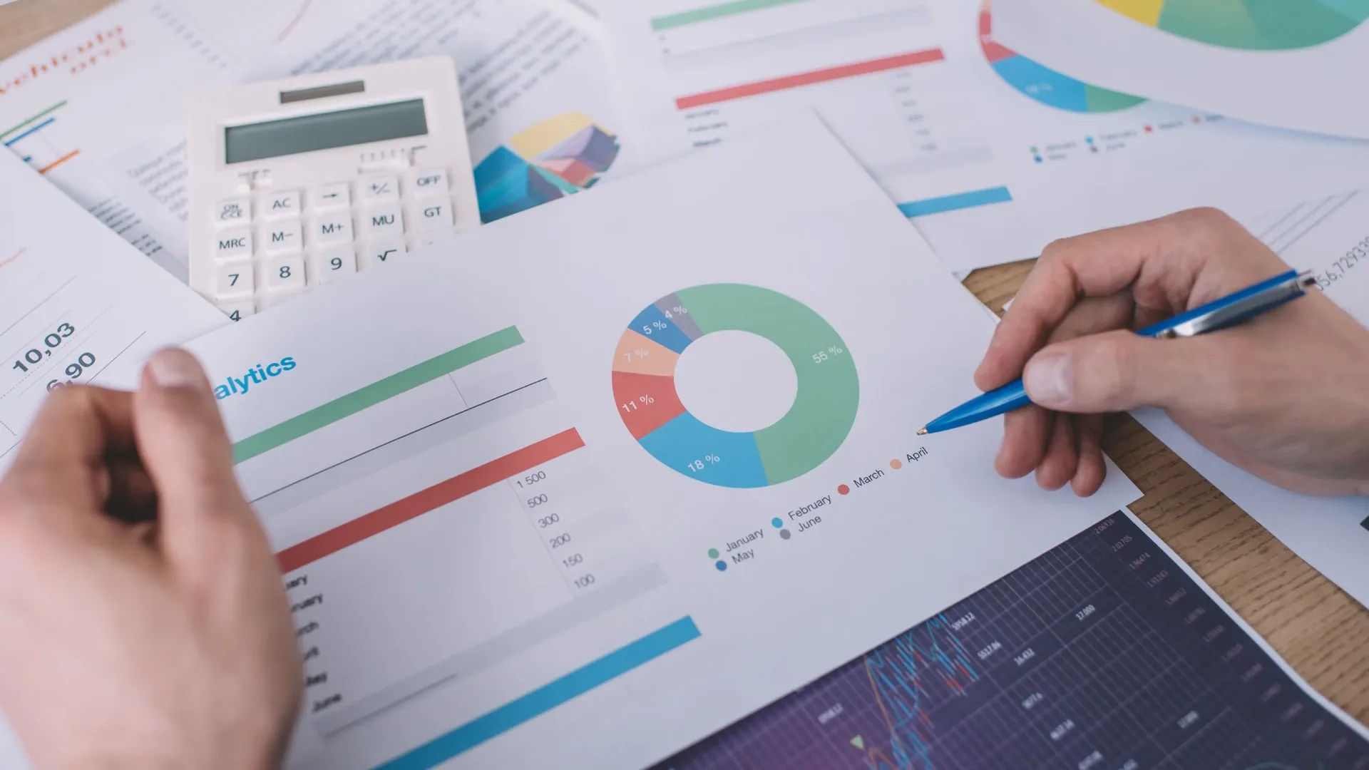 Hands with a pen and a calculator over printed pages of bar charts and donut charts.
