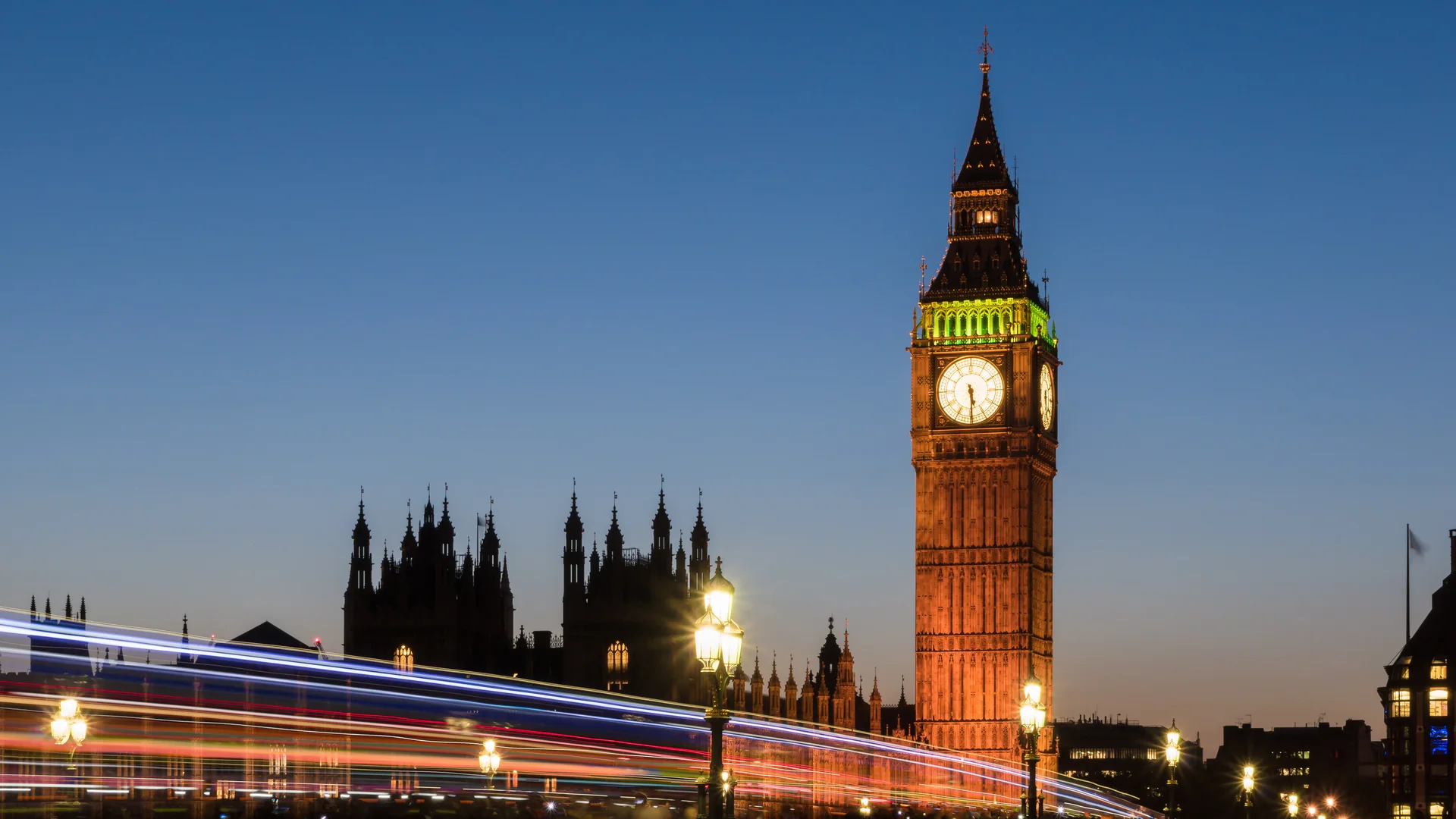 The clock tower of the Palace of Westminster lit at dusk against a deep blue sky, with streaks of light from passing traffic crossing Westminster Bridge and lit lamp posts in the foreground.