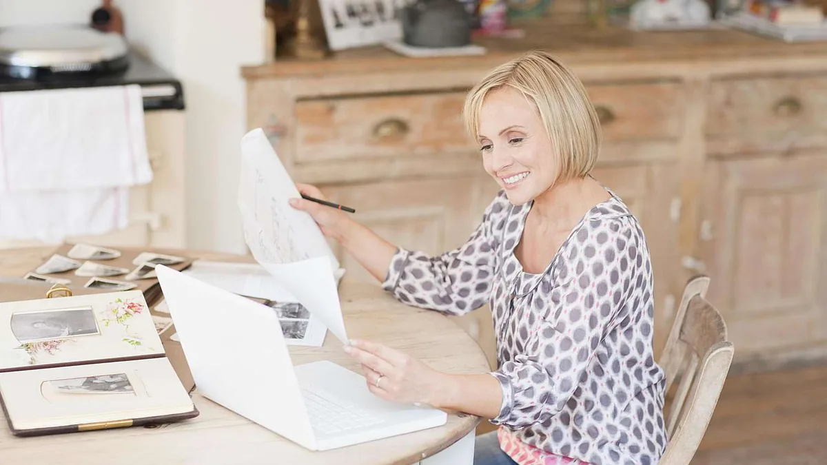 A smiling woman at a kitchen table holds a sheet of paper beside an open laptop and photo albums.