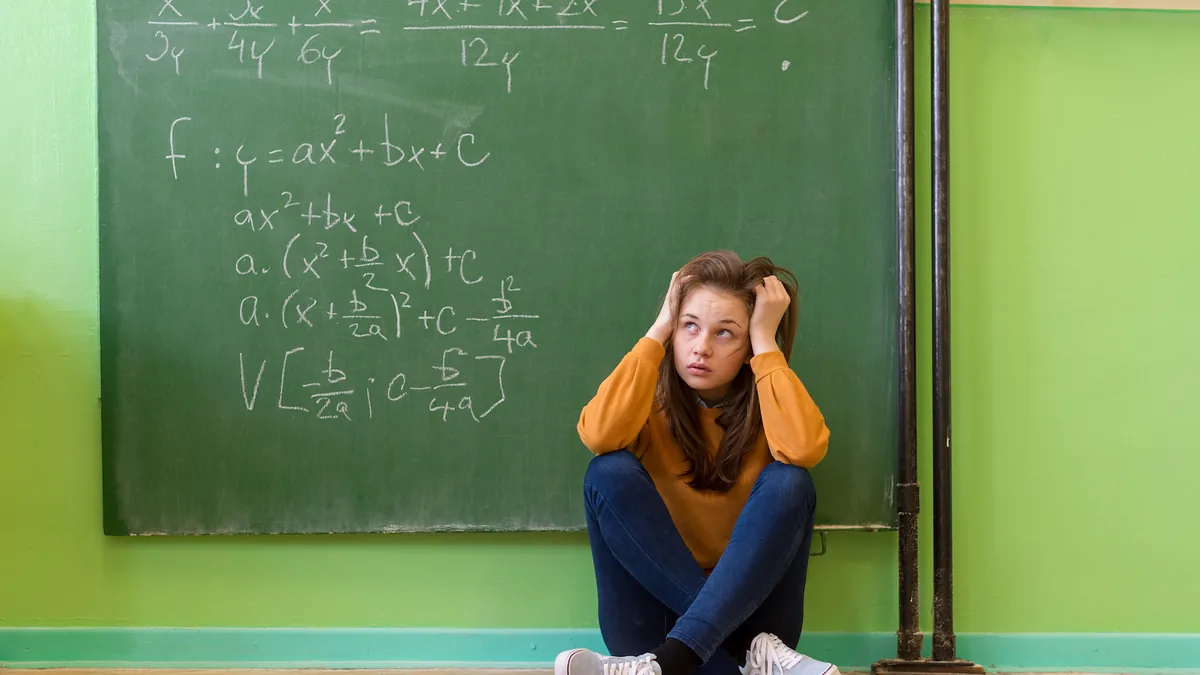 A teenage girl in a yellow top and jeans sits on a classroom floor with both hands in her hair and her eyes turned upward, in front of a green chalkboard covered in algebra equations, against a bright green wall.