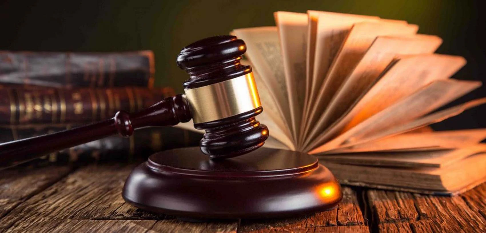A dark wooden gavel with a gold colored band resting on a round wooden block on a wooden table, beside an open book with its pages fanned out and a stack of old leather-bound books.