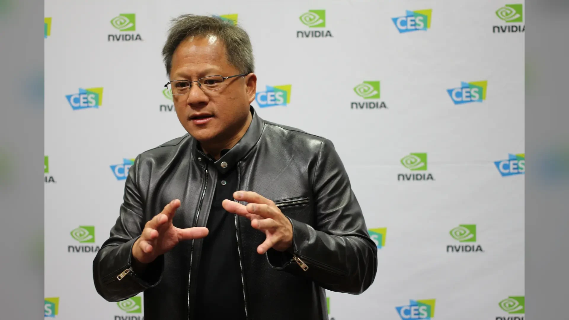 A man with gray hair and glasses, wearing a black leather jacket, gestures with both hands in front of a white backdrop printed with green Nvidia logos and blue CES logos.