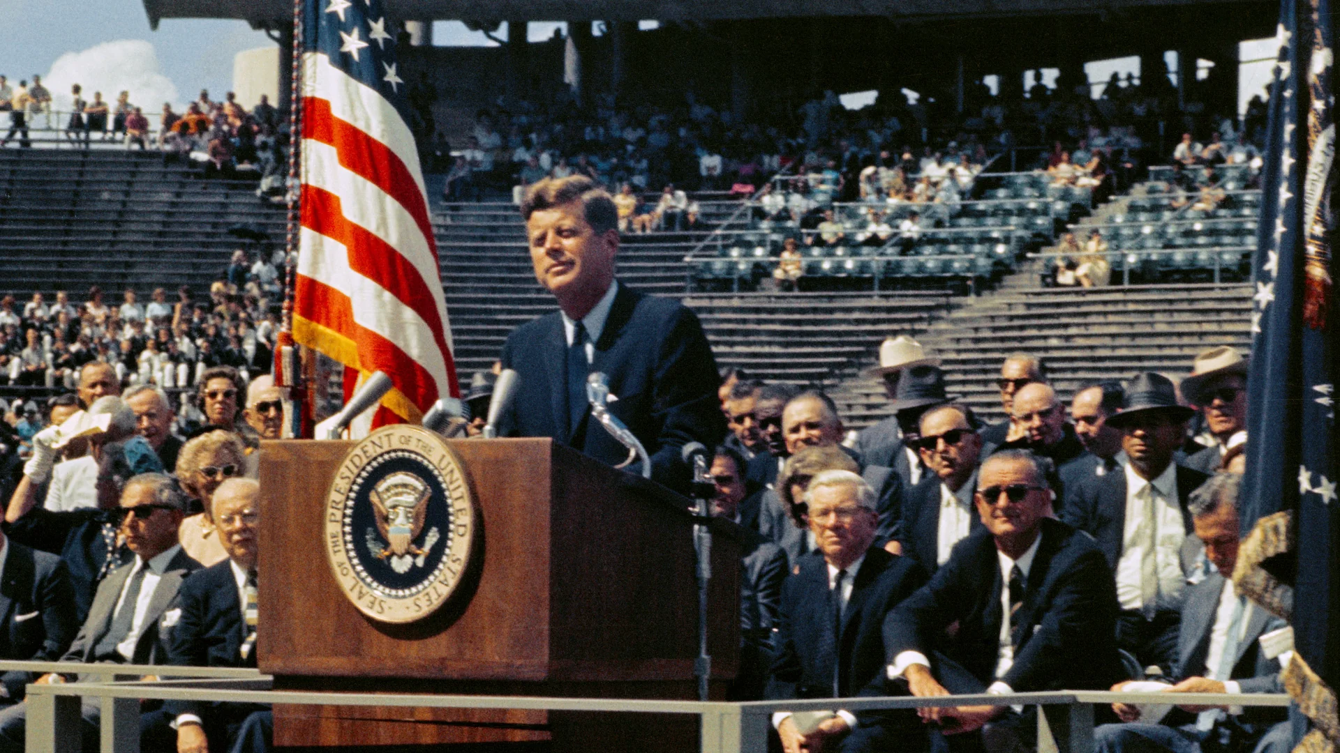 John F. Kennedy speaking at a lectern in a stadium, with an American flag and seated people around him.