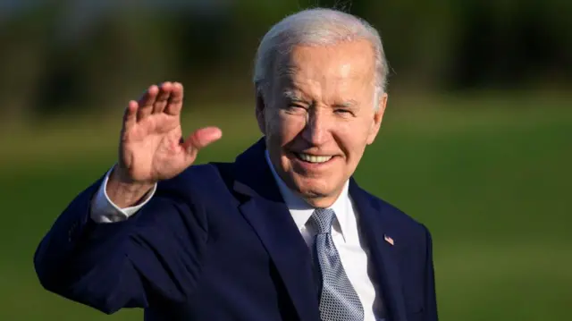 Joe Biden in a dark blue suit and light tie waving with his right hand outdoors against a green lawn.