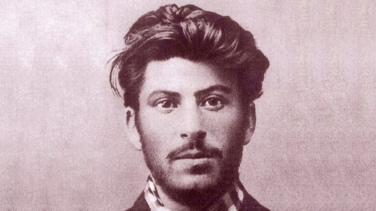 Sepia toned portrait photograph of a young man with thick, wavy dark hair swept back from his forehead, a mustache and a short beard, looking at the camera, with a dark jacket and a patterned collar at the bottom of the frame.