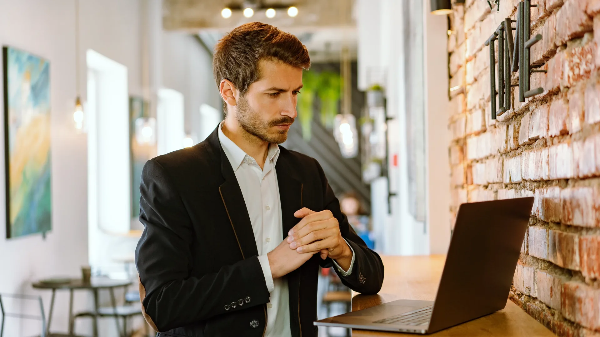 A man in a dark suit jacket stands with clasped hands beside an open laptop in a room with a brick wall.