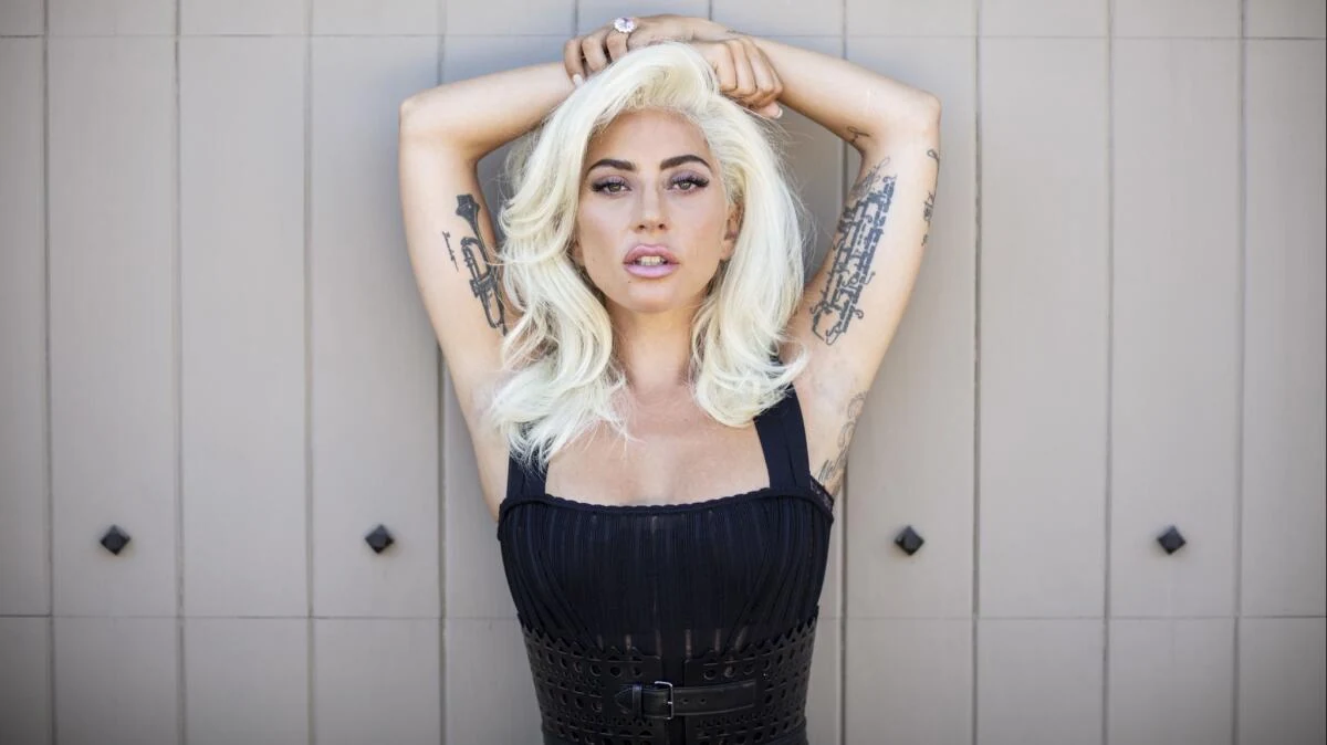 A woman with wavy platinum blond hair stands against a pale paneled wall with both arms raised above her head, wearing a black sleeveless top, with tattoos visible on both forearms.