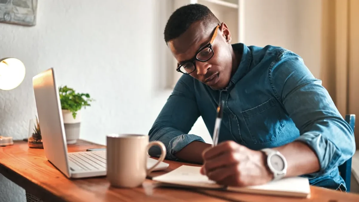 A man with short black hair and dark framed glasses, wearing a blue denim shirt with rolled sleeves and a wristwatch, writes with a pen in a notebook at a wooden desk, with an open laptop, a beige mug and a small potted plant beside him.
