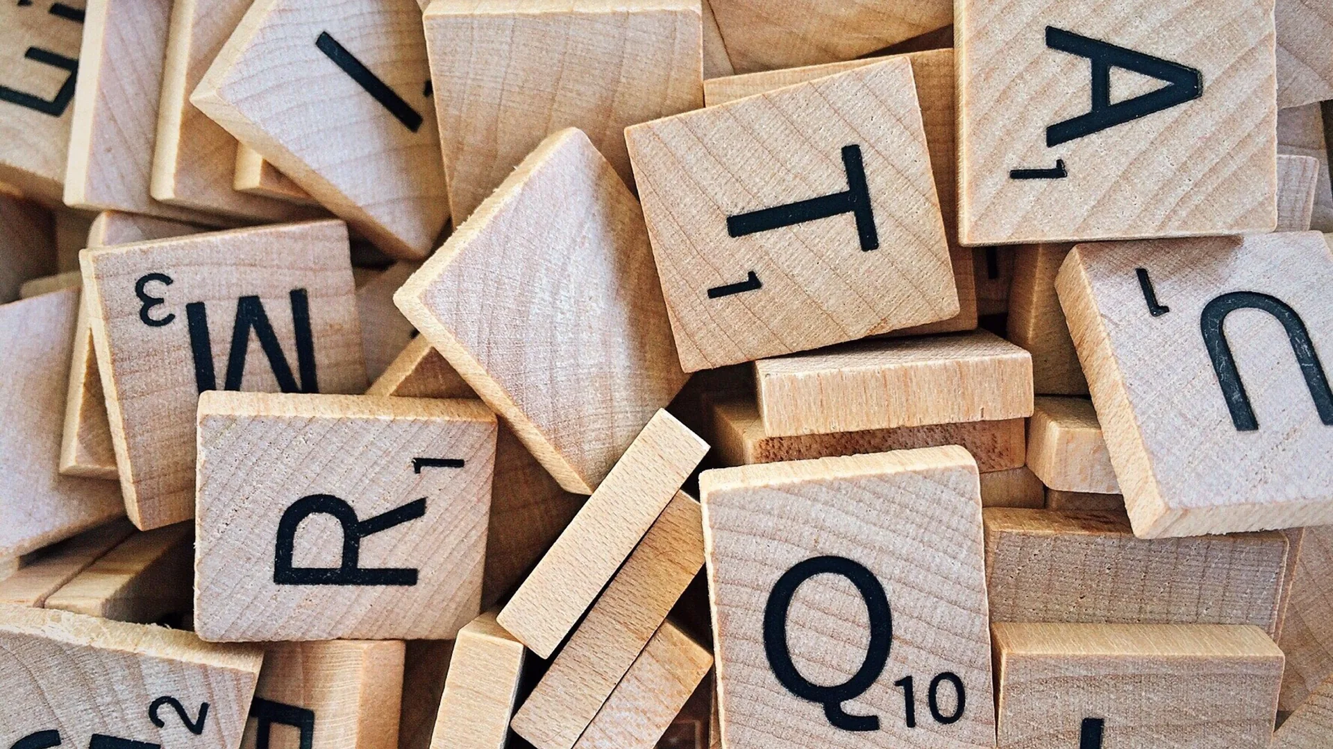 A loose pile of wooden letter tiles, each printed with a letter and a small number, such as Q with 10 and R with 1.