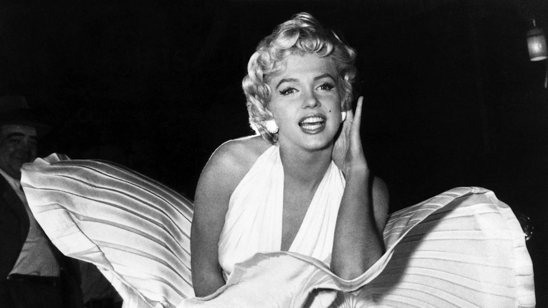 Marilyn Monroe in a white halter dress with a billowing skirt, smiling with one hand beside her face.