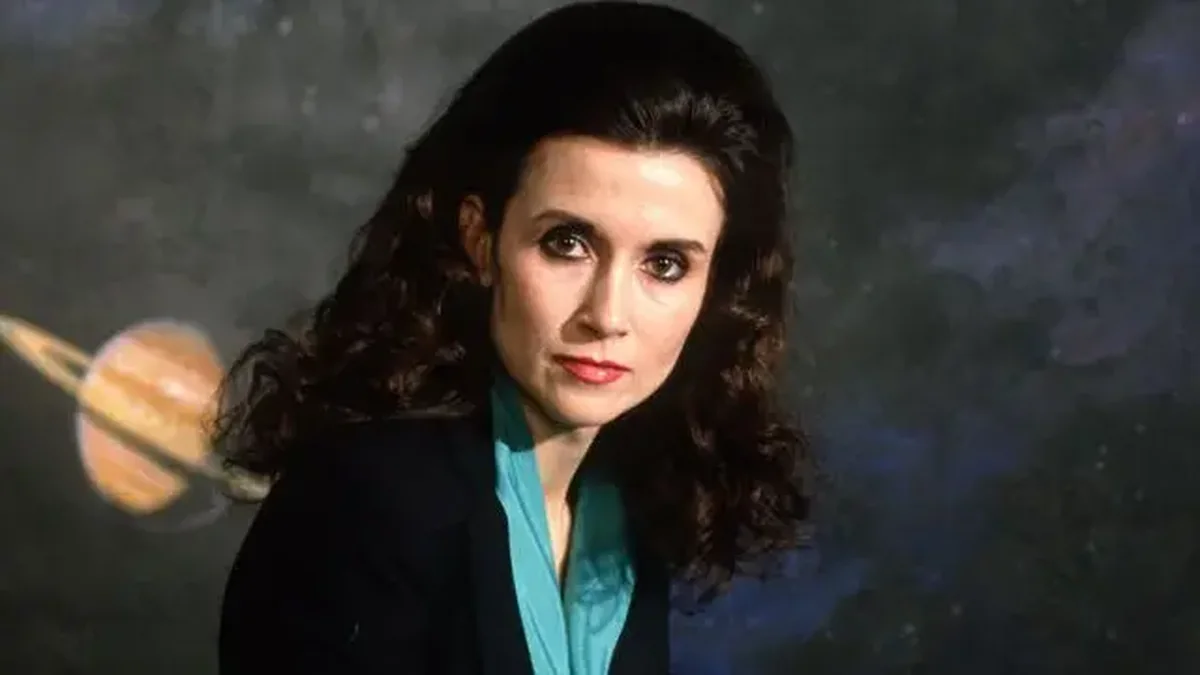A woman with dark hair and a patterned scarf sits at a desk in front of shelves of books, looking toward the camera with a pen in her hand and a stack of letters beside her.