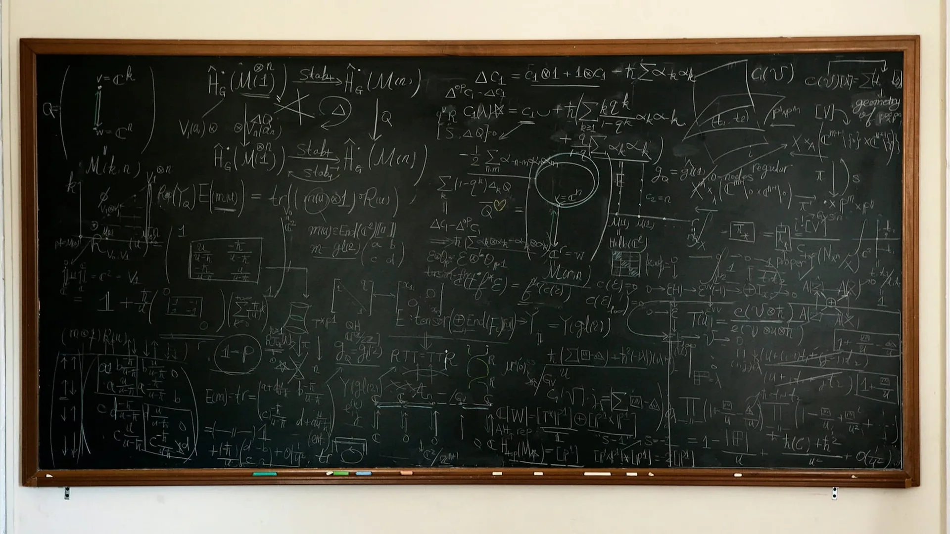 A wooden framed blackboard on a pale wall, covered from edge to edge with handwritten chalk equations, arrows and diagrams, with a few pieces of chalk resting on the ledge below.