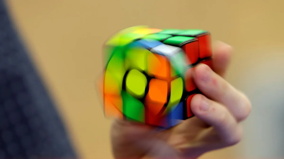 A brightly colored cube puzzle blurred by motion in a hand, against a tan background, with part of a gray sleeve at the left edge.