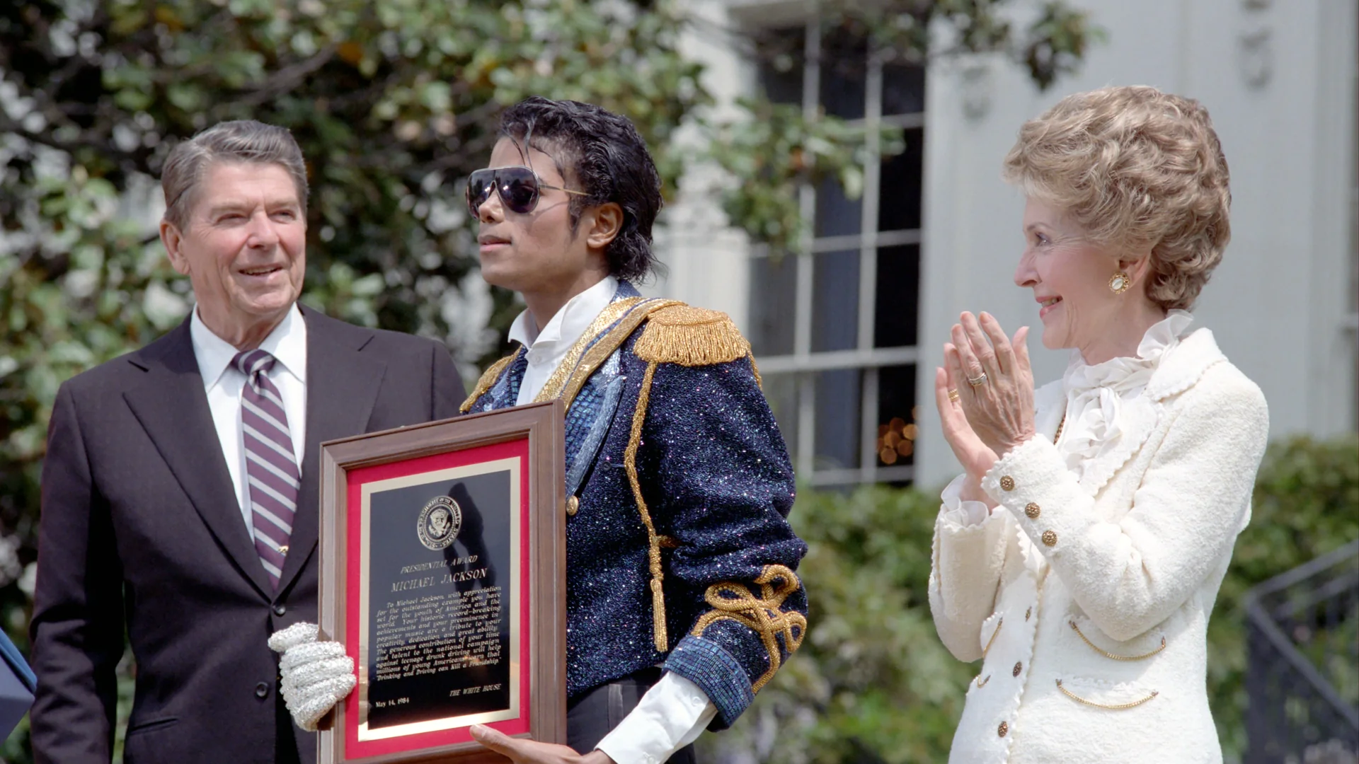Michael Jackson wearing sunglasses and a blue jacket with gold trim, holding a framed award between Ronald and Nancy Reagan.