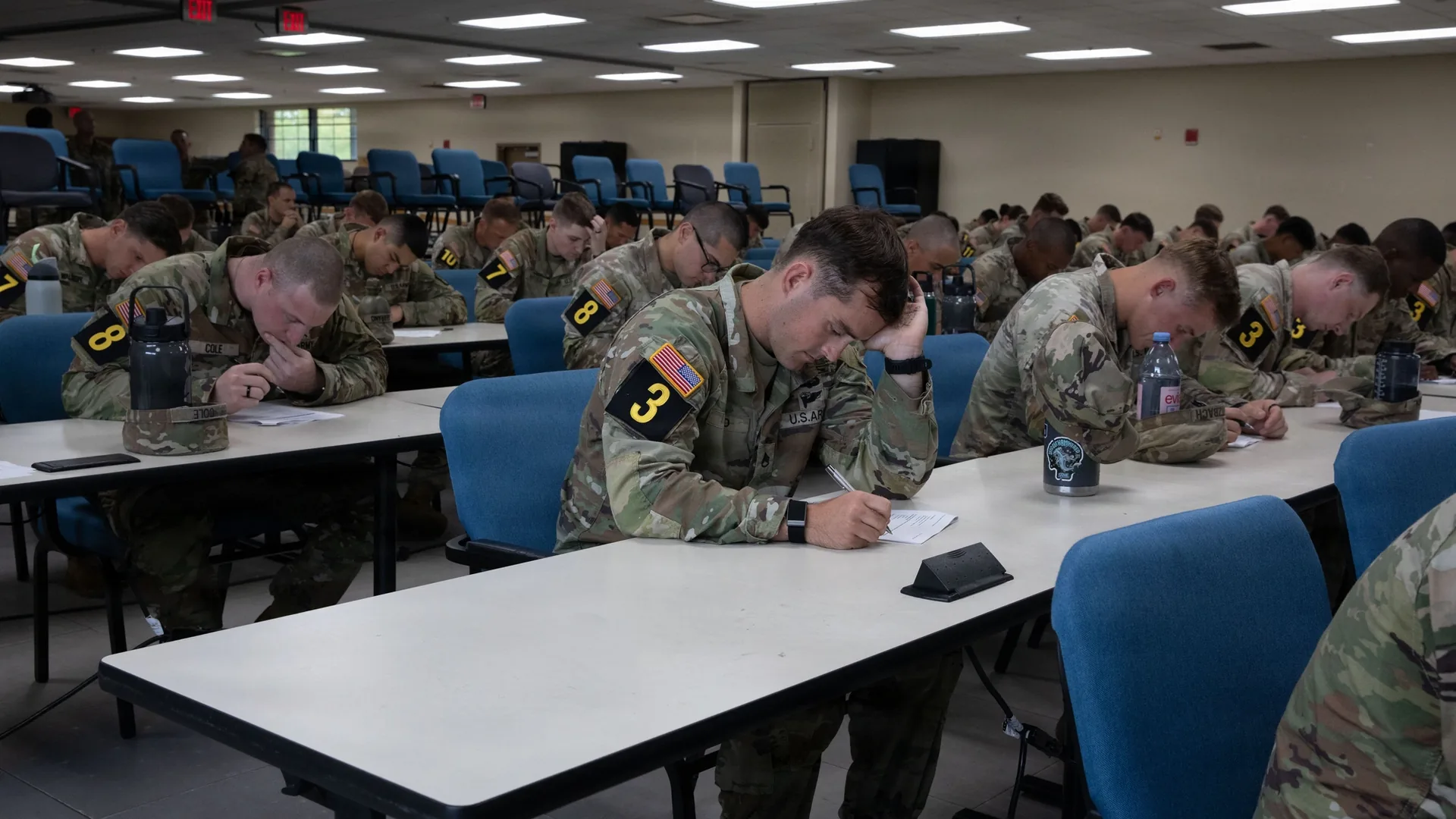 U.S. Army soldiers in camouflage uniforms with numbered armbands write a paper exam at long tables in a large hall.