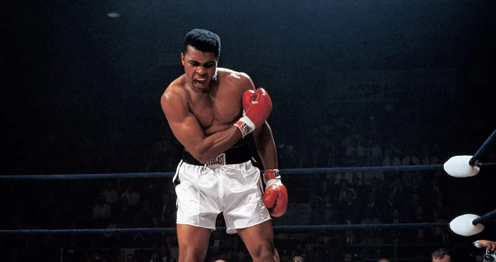 Muhammad Ali standing in a boxing ring in white trunks and red gloves, shouting.