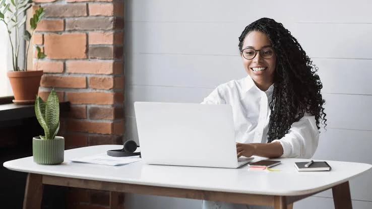 A smiling woman with long dark curly hair and glasses, wearing a white shirt, sits at a white desk and types on an open laptop, with a brick wall, a potted plant, a pair of headphones and a small notebook around her.