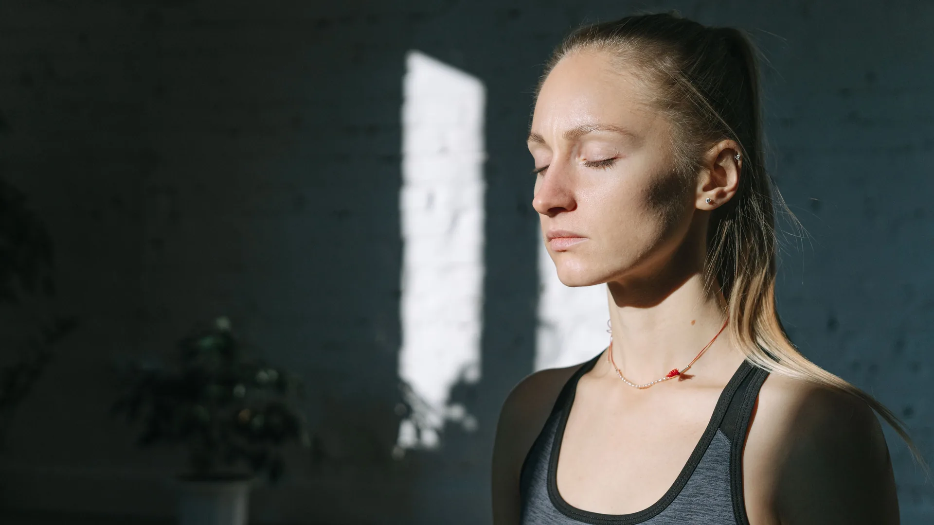 A woman in a sports top stands with her eyes closed in a dim room crossed by a band of sunlight.