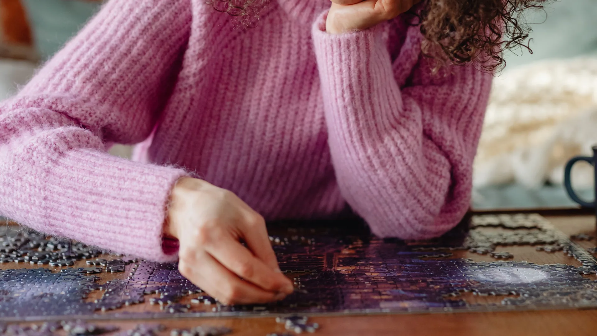A woman in a pink sweater rests her chin on one hand while placing a piece in a jigsaw puzzle.