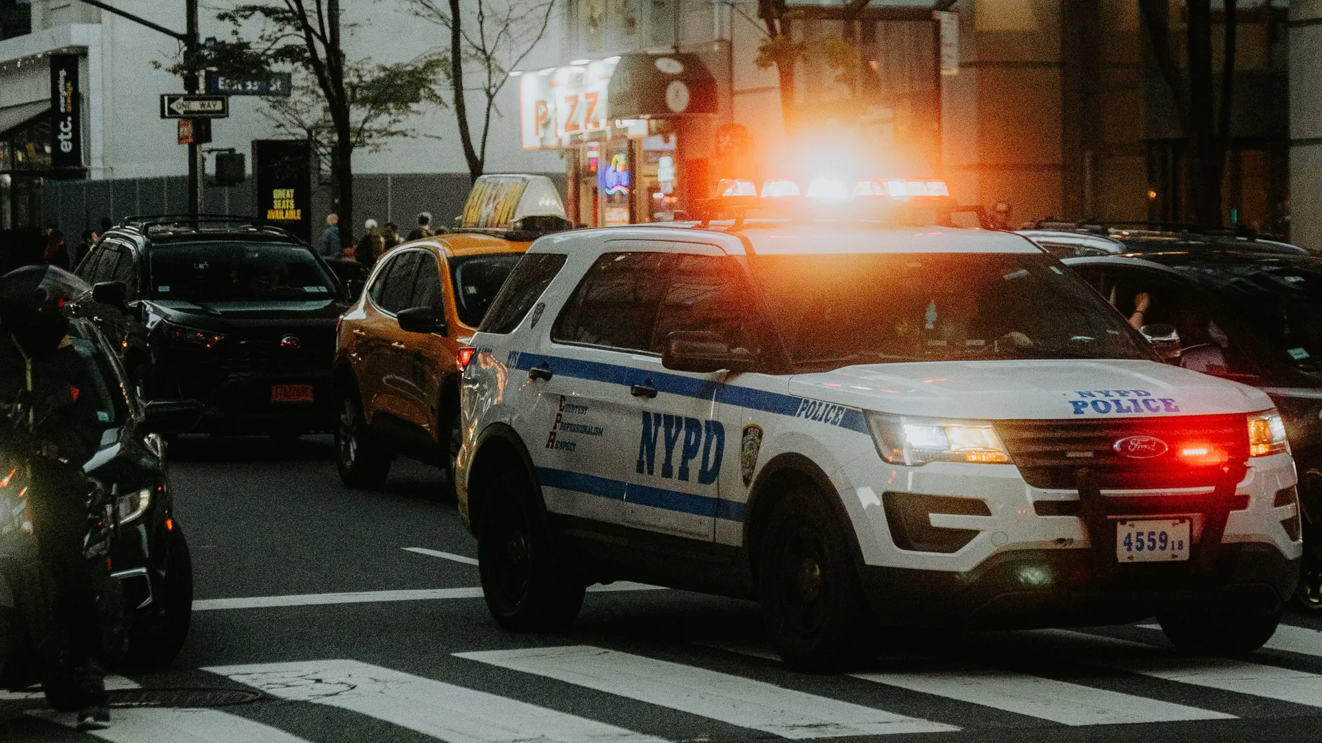 A New York City police SUV with its roof lights on crosses a crosswalk among taxis and cars.