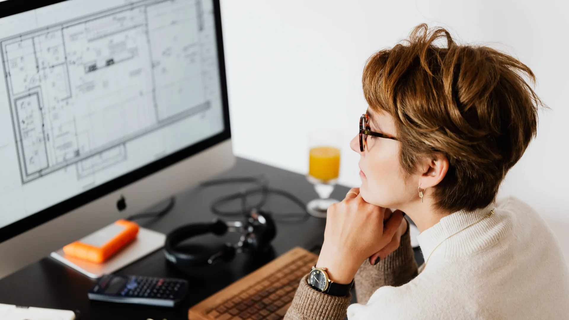 A woman with short hair and glasses rests her chin on her hand while looking at a floor plan on a desktop monitor.