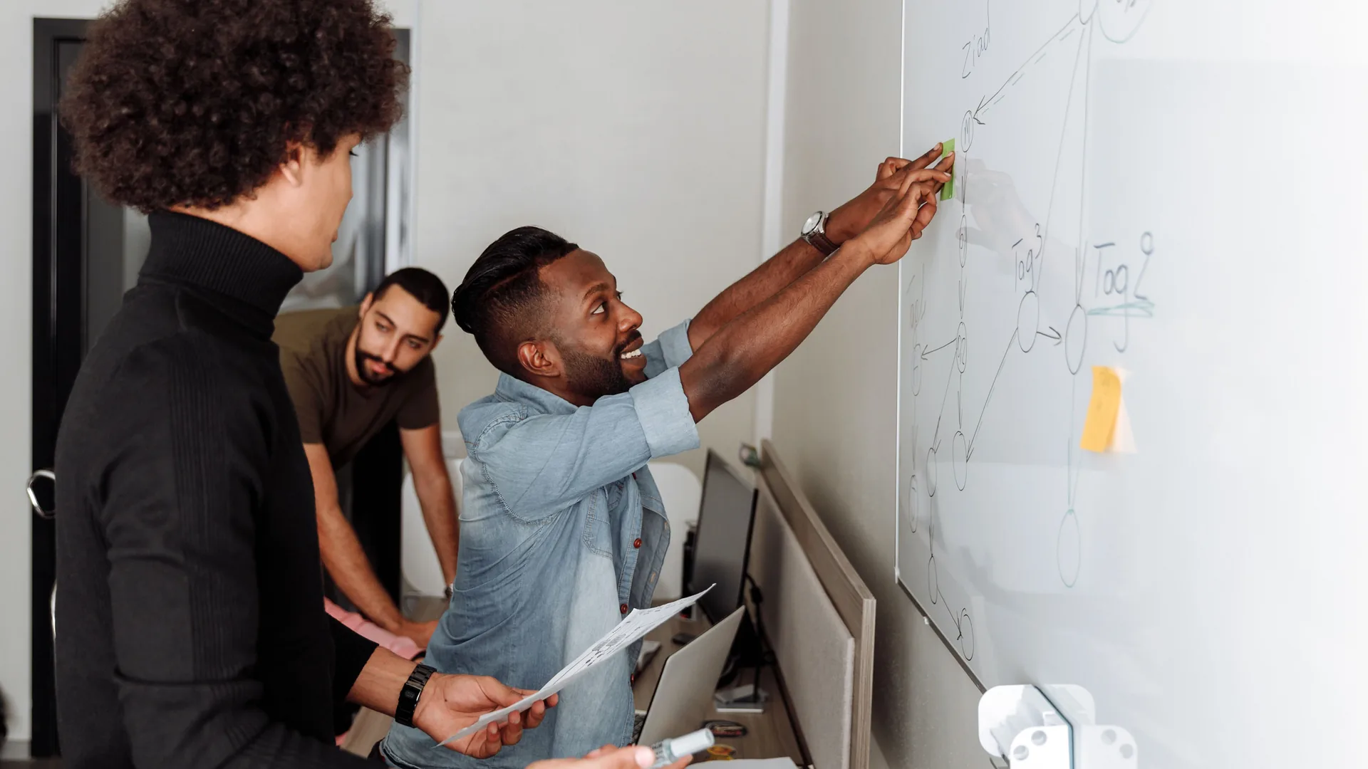 Three men stand at a whiteboard as one reaches up to point at a diagram of circles and arrows.