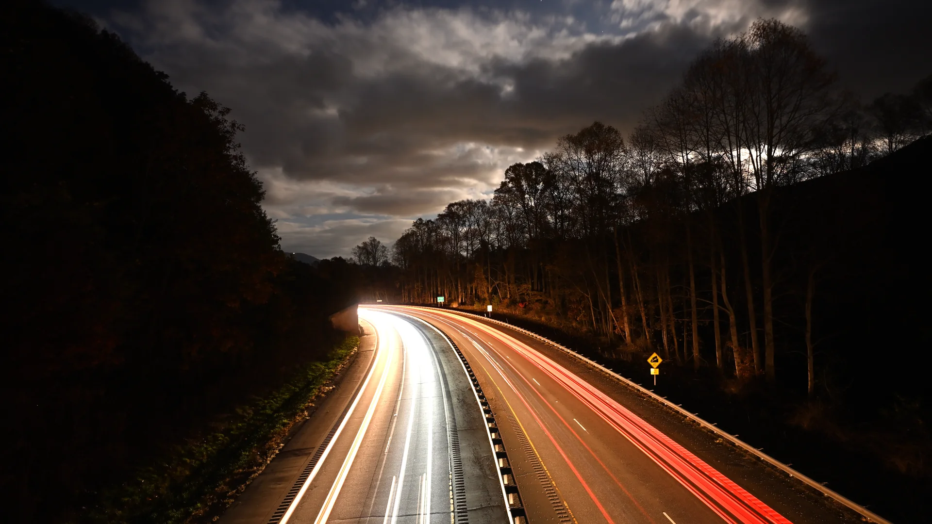 A long exposure night photograph of a divided highway curving through a wooded mountain gap, with white headlight trails on the left roadway and red taillight trails on the right beneath a cloudy sky.