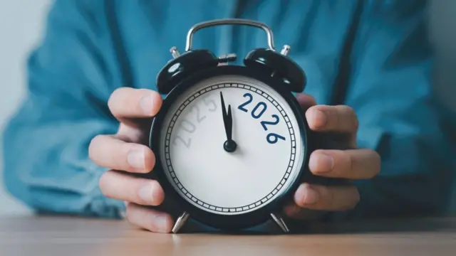 Two hands in a blue sleeve holding a black twin bell alarm clock whose face reads 2026, with the hands near twelve.