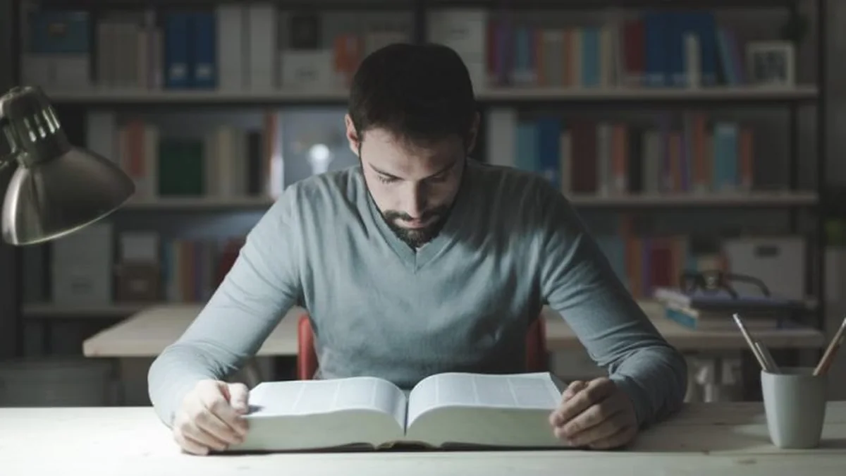 A bearded man in a grey sweater reads a large open book under a desk lamp in a dim library, with shelves of books blurred behind him.
