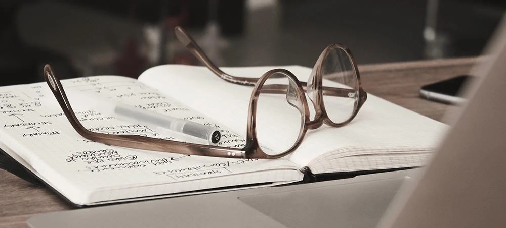 An open lined notebook with handwritten notes and small diagrams lies on a wooden desk, with a pair of brown tortoiseshell glasses resting across its pages, a silver pen with its cap beside them, what looks like a smartphone at the upper right and the edge of a laptop in the foreground.