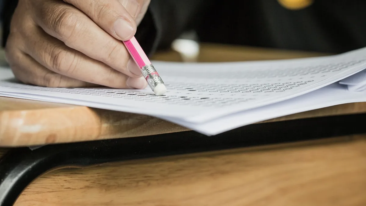 A hand holding a pink pencil fills in a bubble answer sheet on a wooden desk.