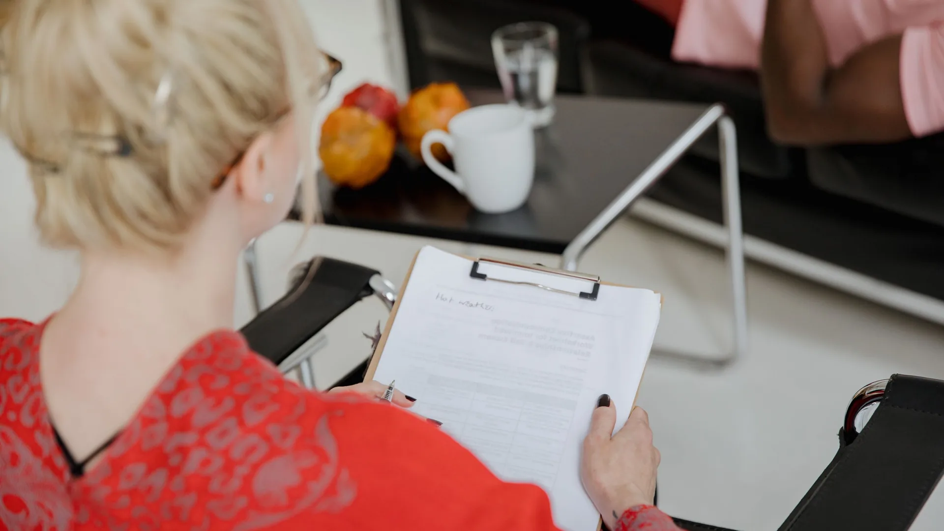A woman in a red top, seen from behind, holds a clipboard with a form during a session.