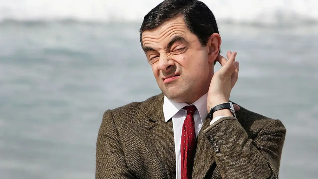 Rowan Atkinson pulling a comic face with a hand at his ear, in a tweed jacket.