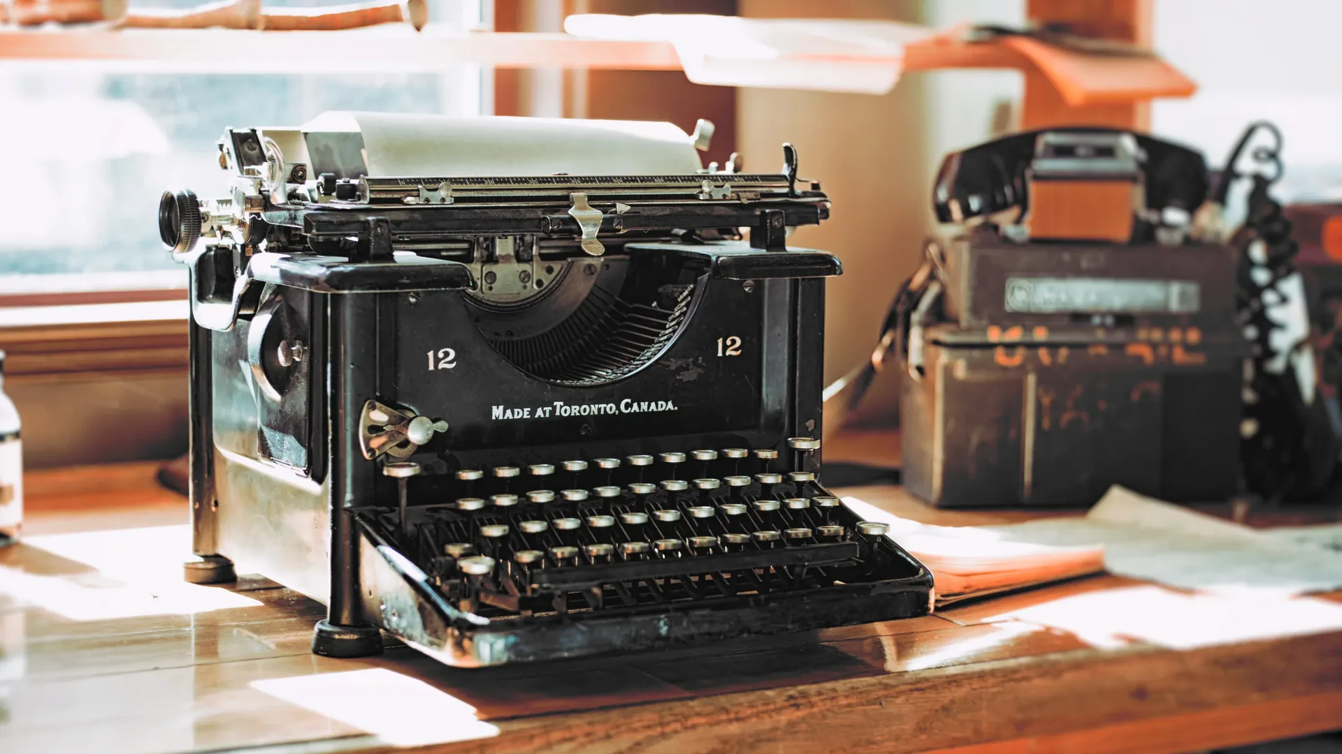 A black vintage typewriter marked Made at Toronto, Canada, with a sheet of paper loaded, on a wooden desk by a window.