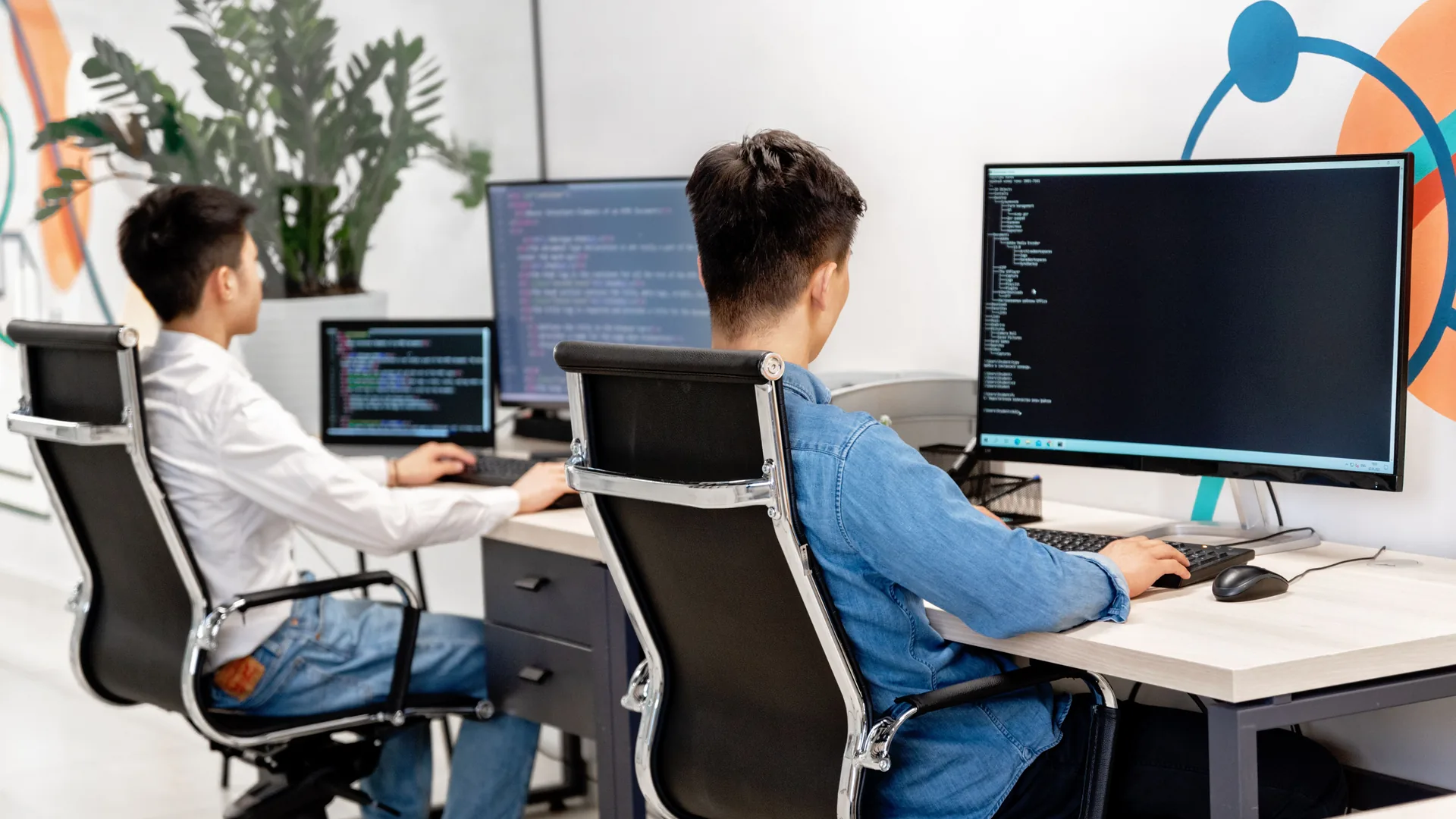 Two men sit at separate desks in an office, facing computer monitors with lines of code on dark screens.