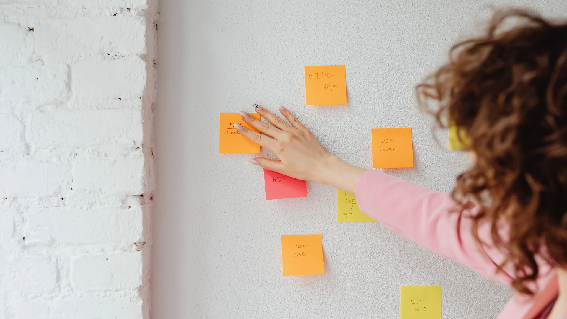 A woman with curly hair, seen from behind, presses a sticky note onto a white wall dotted with notes.