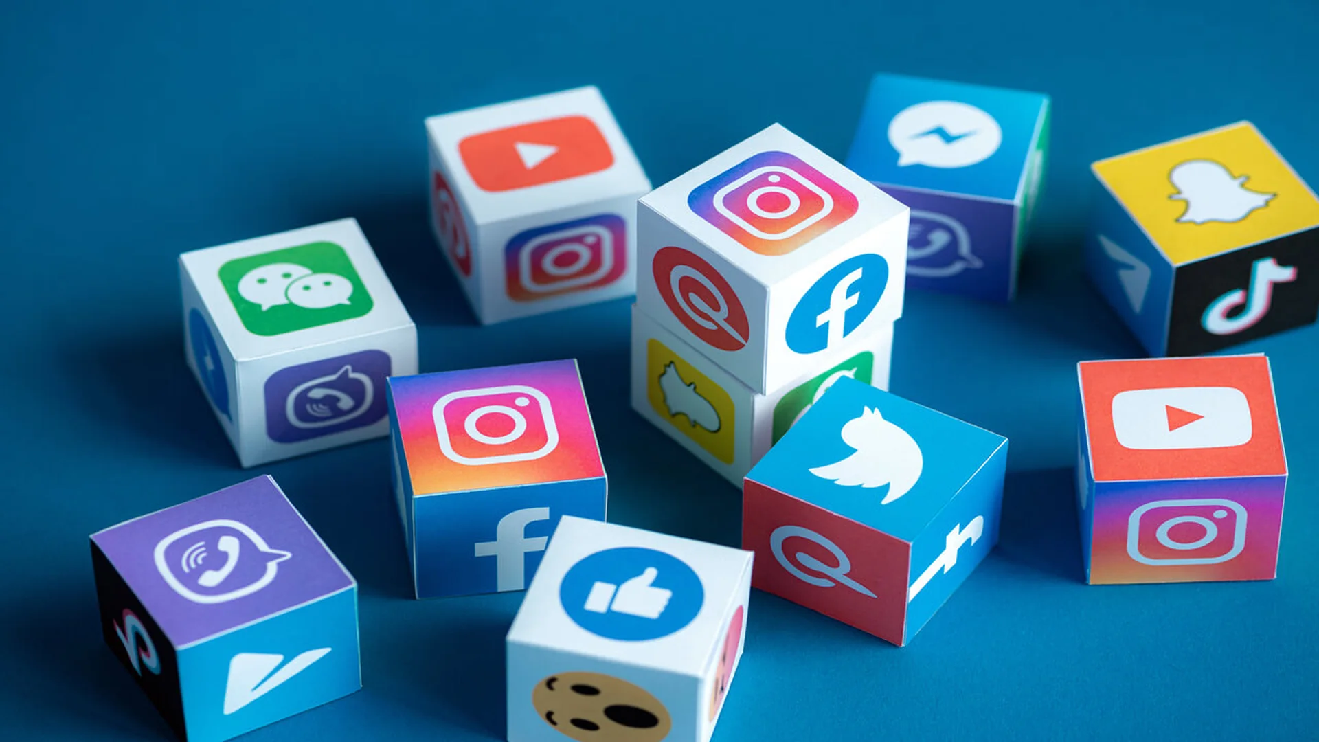 Paper cubes printed with the icons of social media and messaging apps, including Instagram, Facebook, YouTube, Snapchat, TikTok, WeChat and a bird logo, scattered on a blue surface.